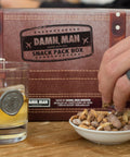 Damn, Man Snacks Snack Pack Box on table with a hand reaching in to grab a handful of nuts in a small bowl and a glass of whiskey on the side
